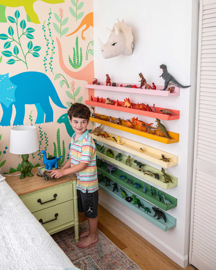 Young boy playing with toys in a bedroom. Dinosaur-themed wallpaper on the wall, and shelves of rainbow-colored dinosaur toys behind him
