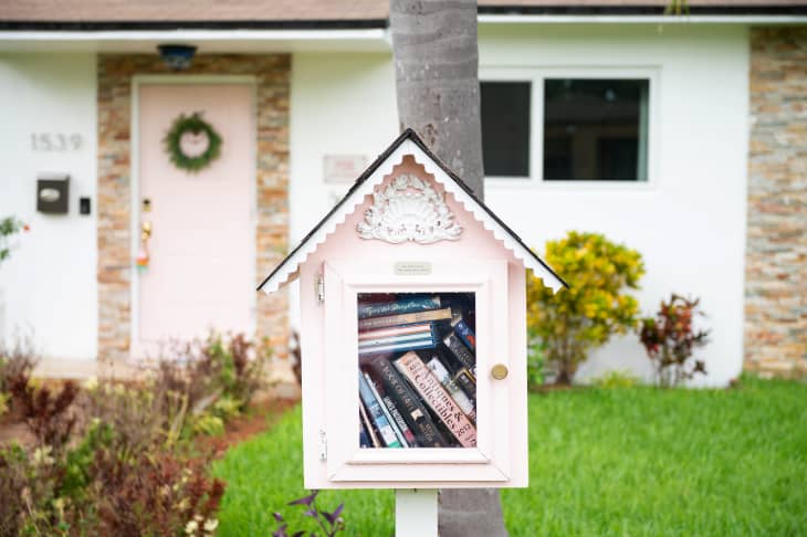 little pink house-shaped free library full of books posted on tree trunk in front of white and pink house. Green lawn and some bushes around