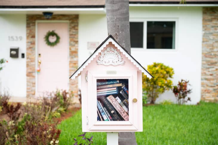 little free library in the shape of a pink house full of books placed on a tree in front of a white and pink house.  A green lawn and some bushes around