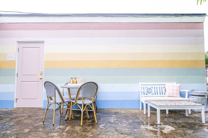patio of house with pastel rainbow striped wall. a table with chairs and a white bench with a pink and white pillow
