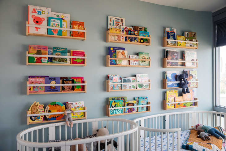 Kids' room in blue with 2 cribs and shelves full of front facing kids' books on the wall behind