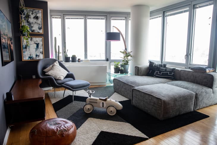 living or family room with a gray sectional sofa, a gray/blue armchair with a footstool and throw pillows, a black and gray graphic rug, a kids' ride-on scooter toy. There are large windows and artwork on the walls