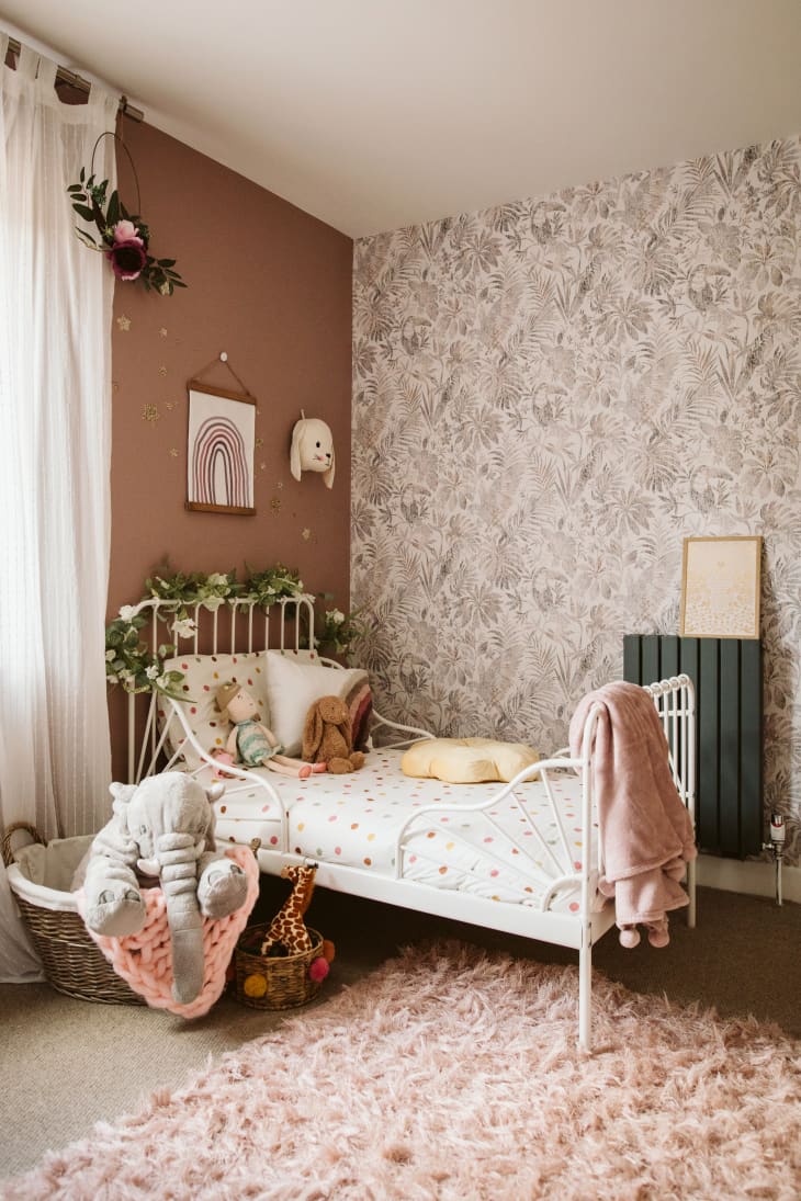Photo of kid's bedroom, focus is on the white iron-framed bed. There is blush-colored shag carpet and decorative wallpaper