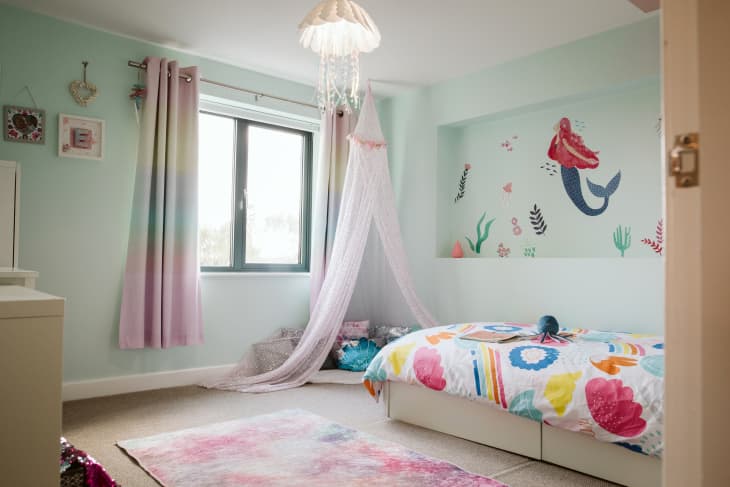 Kid's room with mermaid theme. Light aqua colored walls, mermaid mural on one wall. White jellyfish hanging lamp, colorful bedding