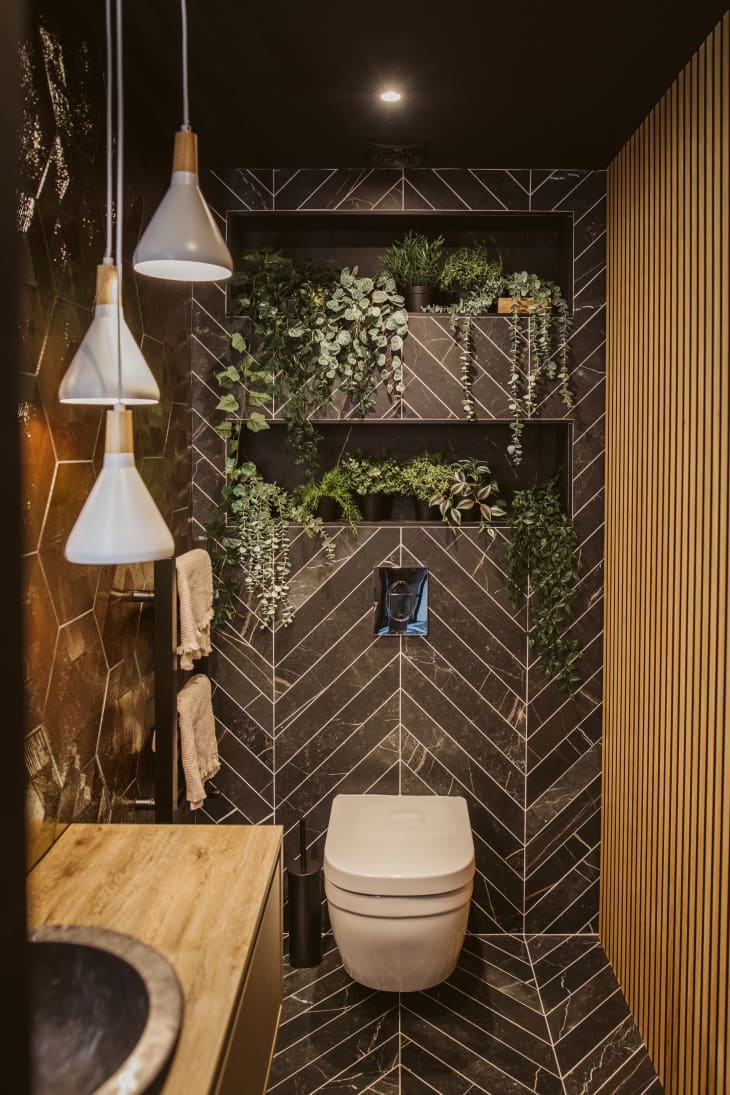 Vertical photo of brown tile bathroom. Tiles are in a chevron shape. There are 3 hanging pendant lights and a wood counter. Lots of plants on shelves behind the toilet