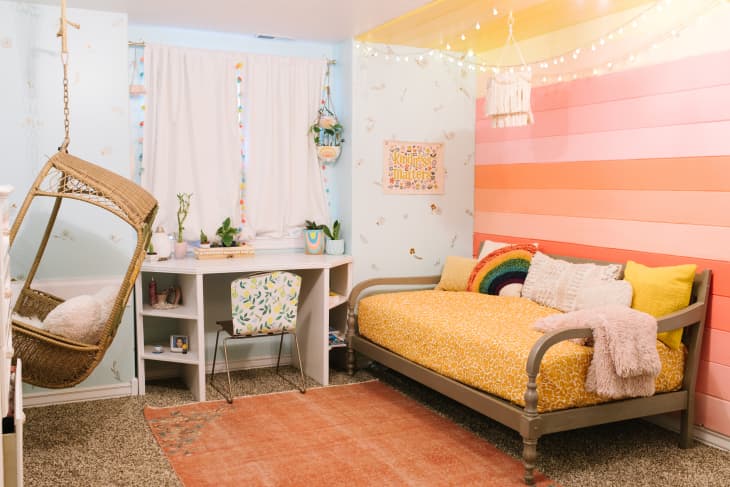 Kids' room with pastel orange pink and yellow rainbow wall and wicker hanging swing.