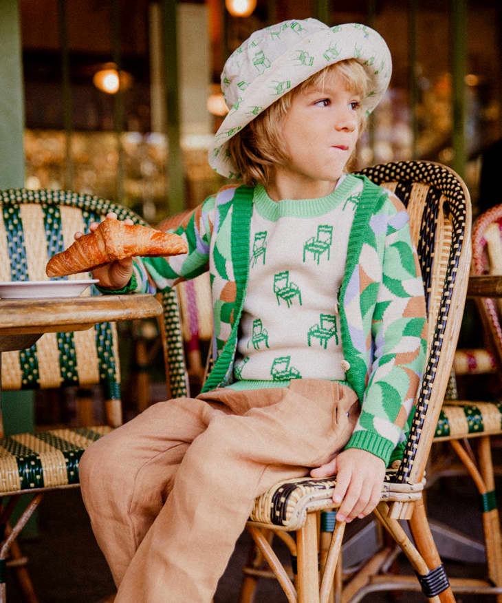 Boy with bucket hat and green patttern Oeuf sweater sitting at table with croissant