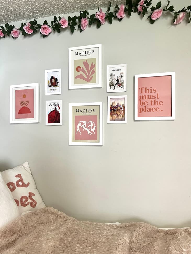 Dorm room with pale green walls, bed with pink and white bedding and throw pillows, faux pink roses and leaves hanging down walls, over bed, gallery wall by bed of pink and white art