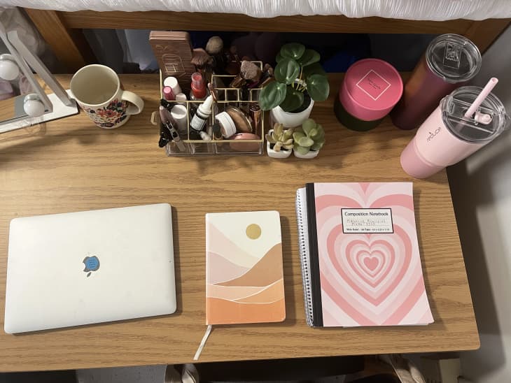 Dorm room with pale green walls, bed with pink and white bedding and throw pillows, faux pink roses and leaves hanging down walls, over bed, gallery wall by bed of pink and white art. Detail of desk area with laptop, sunset graphic notebook/journal, comp book with pink hearts, personal items around