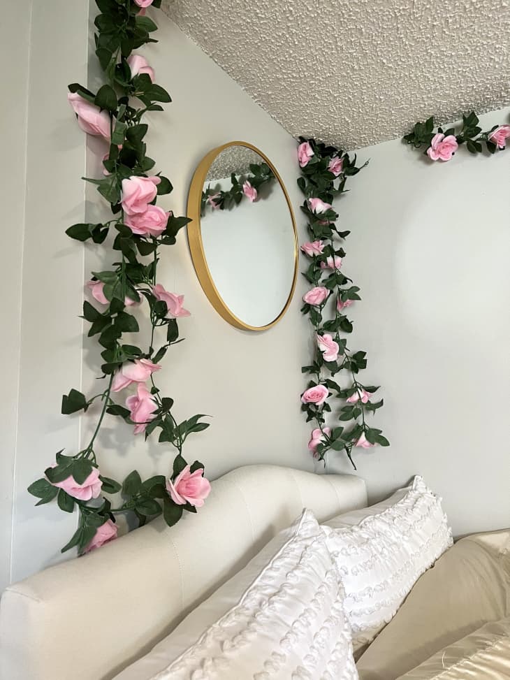 Dorm room with pale green walls, bed with pink and white bedding and throw pillows, faux pink roses and leaves hanging down walls, over bed, gallery wall by bed of pink and white art