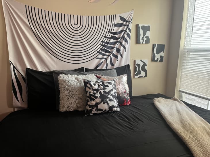black sheets, black and white curved line tapestry above bed, black and white squiggly art, beige throw blanket, black and white throw pillows, window, blinds
