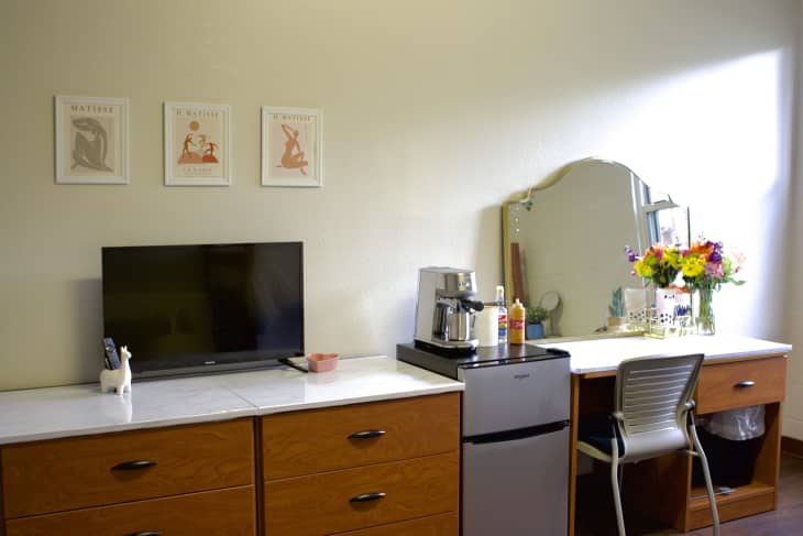A tv on dressers in a dorm room next to a desk and coffee machine.