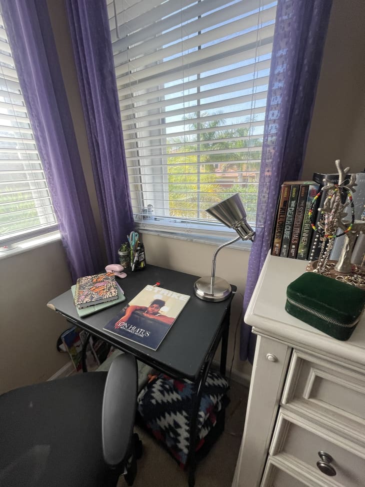 dorm room detail of small black desk in front of window with purple curtains, white dresser with TV, personal grooming items and decor on top of dresser