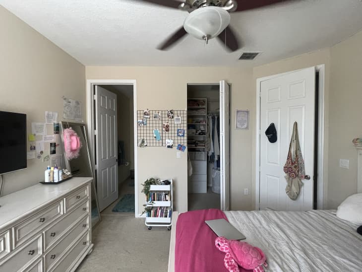 View of one side of dorm room with 2 doors open to the bathroom, and closet. wall grid with photos on it, small organizer cart on floor, dresser to the left of the photo with tv