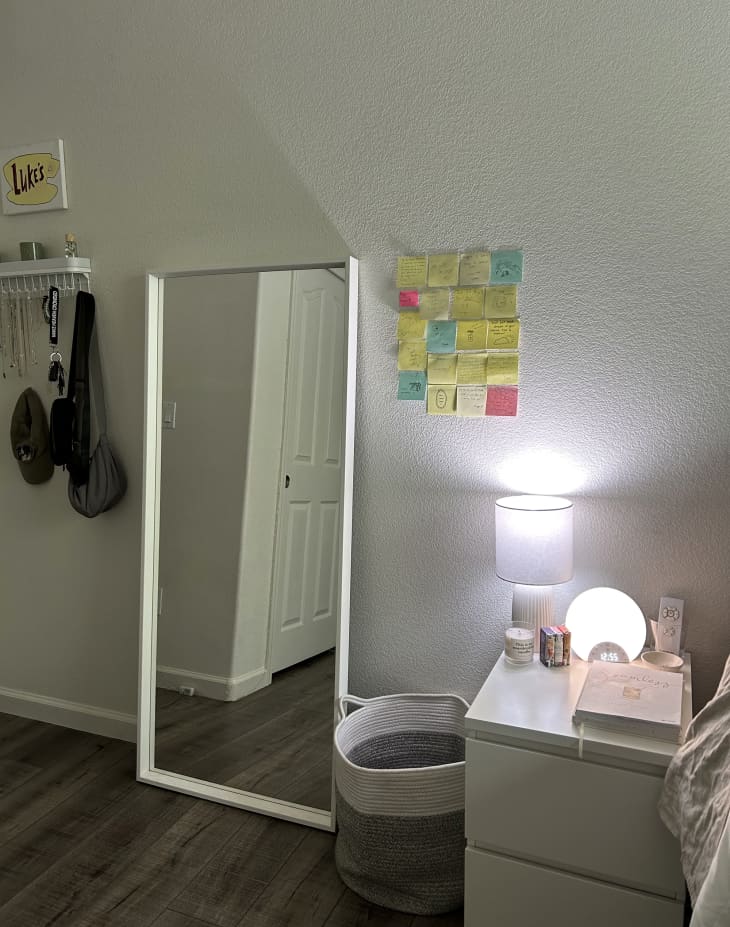 Dorm room with white walls, white frame full length mirror leaning on wall, white bedside table/drawers with lamps, candles, wall hooks with purses and jewelry