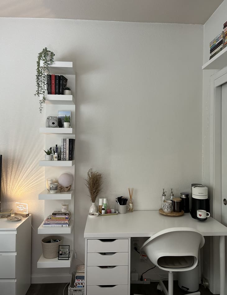 white desk with modern white chair, tall narrow white shelves with books, plant, objects, white walls, in dorm room