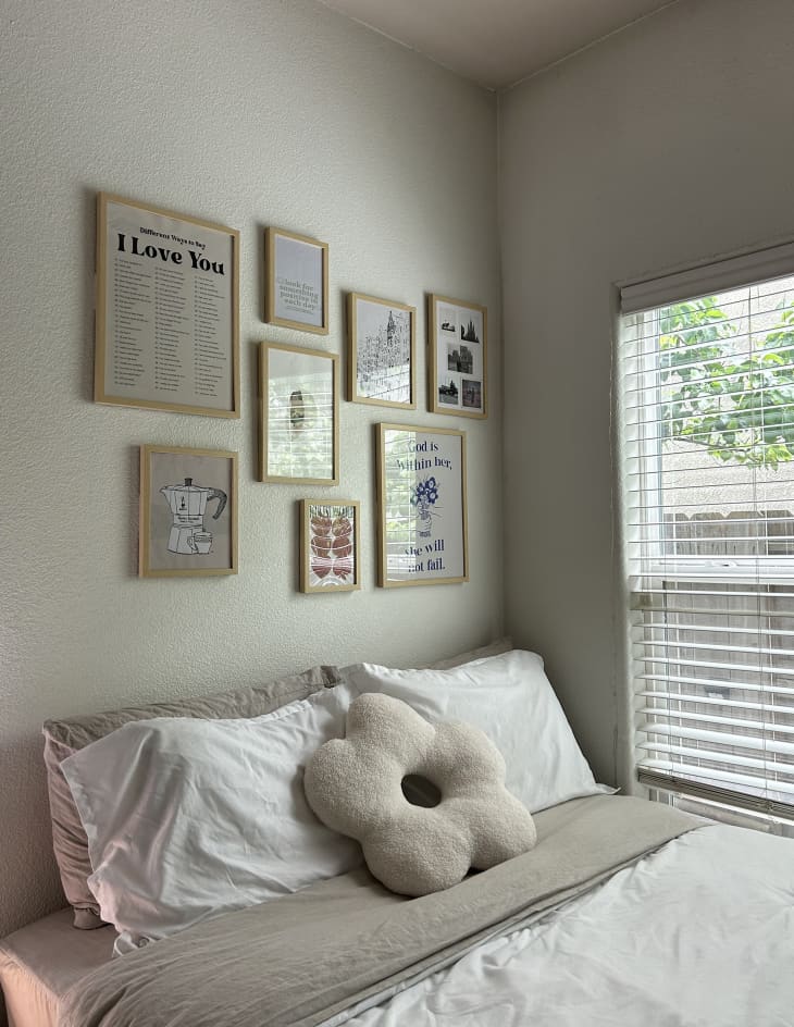 white dorm room with bed with white and gray bedding, plush flower pillow, large window, gallery wall above bed