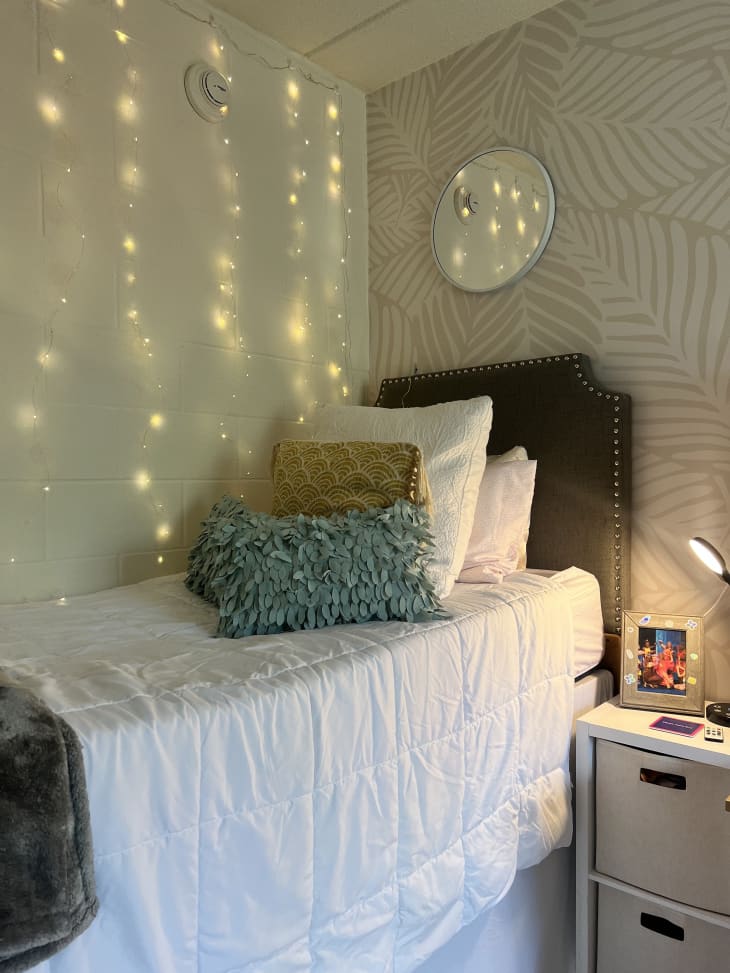 raised bed with white bedding and string lights hanging down wall, round mirror above bed, leaf wallpaper on wall behind bed
