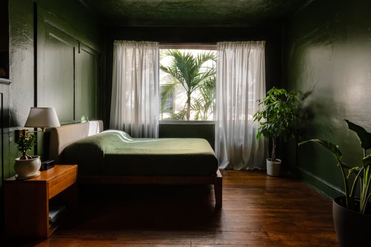 green walls and ceiling, green linens on bed, beige fabric headboard, sheer white curtains, floor plants, wainscoting on walls, wood night table, wall sconces, wood floors