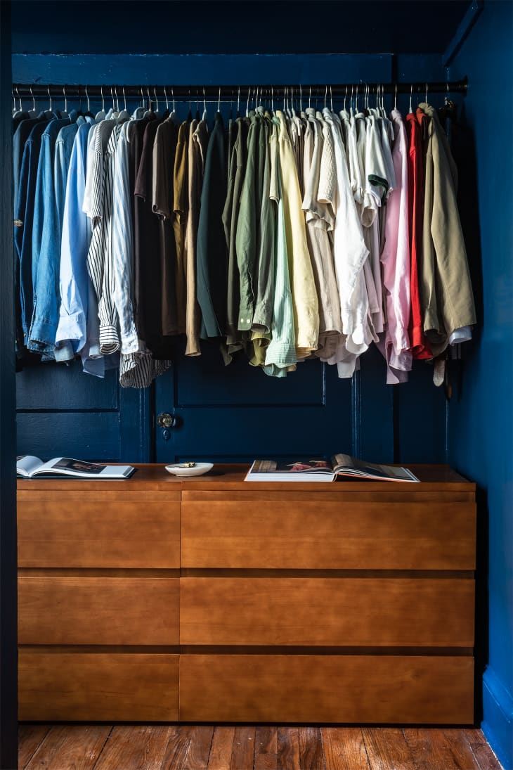 color coded hanging clothes on rack with wood dresser underneath, dark blue walls and wood floors
