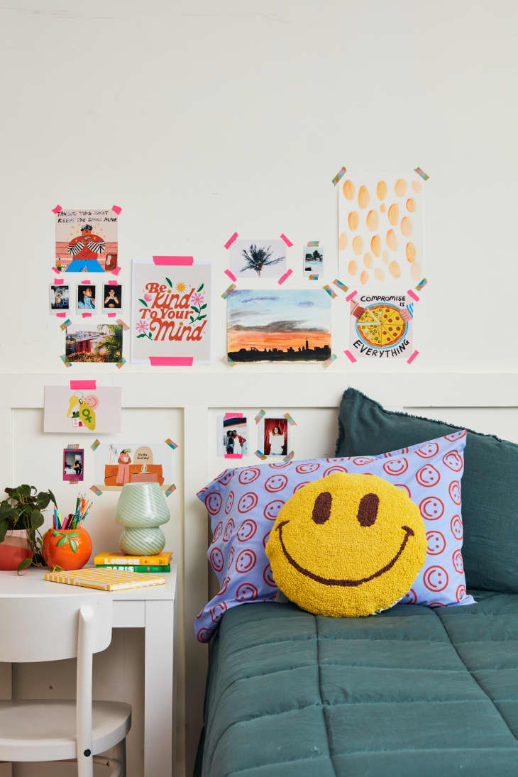 wide shot of a bed with teal sheets, blue pillowcase with red smiley faces on it, a smiley face pillow and a white desk with a green lam, planters, and supplies on top of desk, and a wall with different photos and art
