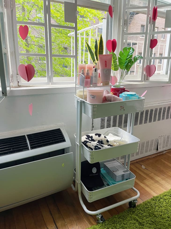 dorm room featuring Amazon Basics 3-Tier Rolling Utility or Kitchen Cart. Room has bed with pink bedding, speckled wallpaper wall with wavy neon, one lavender wall, windows. Cart is in front of a window