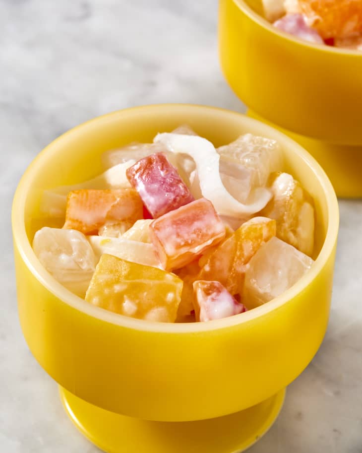 Side view of Filipino fruit salad with chunks of tropical fruit, coconut and condensed milk in a small yellow dish on a marble background with another dish in the background
