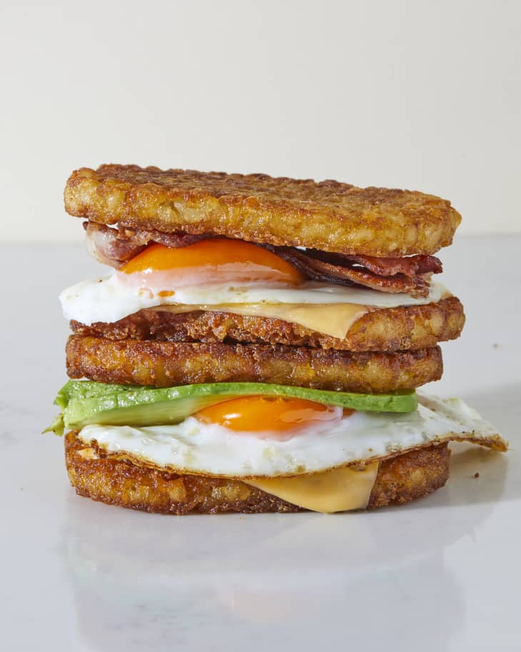 A double stack of hashbrown breakfast sandwiches on a marble surface. The top is loaded with an egg, bacon, and american cheese. The bottom has avocado, egg, and american cheese.