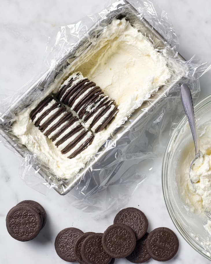 photo showing the process of adding whipped cream and Oreo thins into a loaf pan in rows to make an icebox cake with cookies on the surface and a bowl of whipped cream with a spoon in it to the side