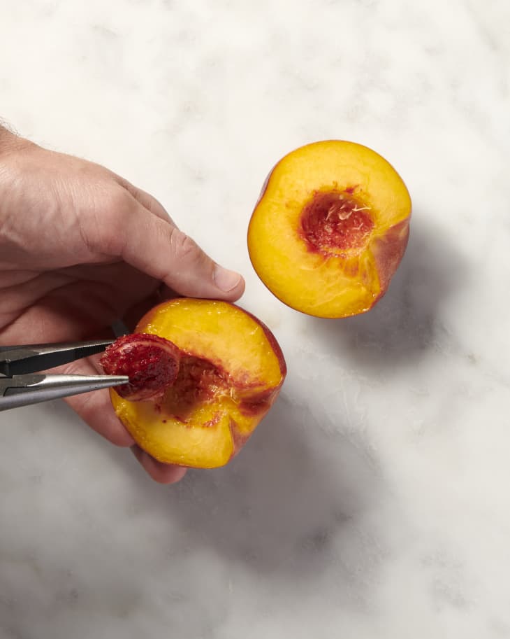 a halved peach with the pit being removed with a pair of needle nose pliers