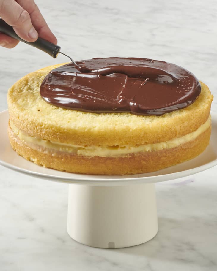 a whole boston cream pie on a cake stand being topped with chocolate ganache