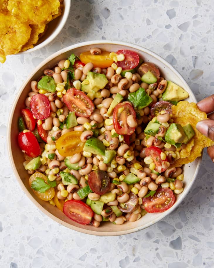 a semi wide shot of a bowl of black eyed pea salad on a Terrazzo background with a hand using a plantain chip to scoop out of the bowl; as a bowl of plantain chips sit besides it