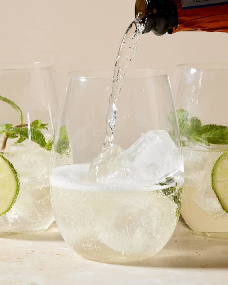a semi wide shot of 3 glass cups of hugo cocktail with limes and mint in each that's on rustic sandy colored surface; a amber colored bottle pours into the cup closest to the camera