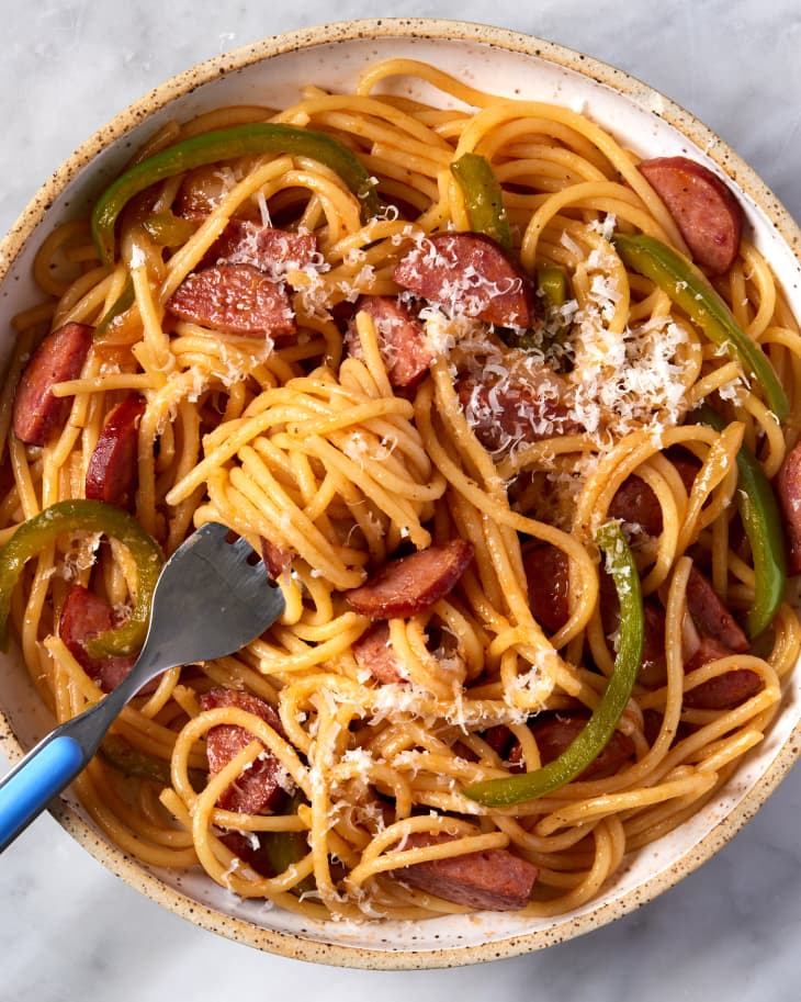 close up overhead shot of bowl with napolitan spaghetti, bell peppers, and sausage with cheese sprinkled on top on a marble surface; a blue fork lays in bowl with spaghetti swirled around it