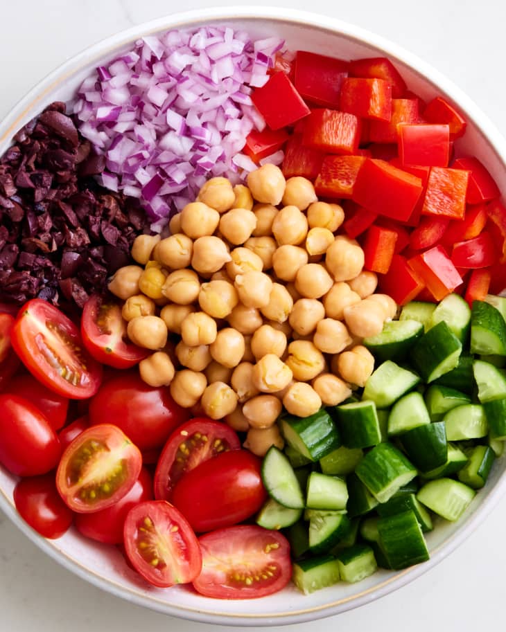 photo of a bowl with chopped olives, red onions, red bell pepper,cucumber, and halved grape tomatoes around a pile of cooked chickpeas on a marble surface