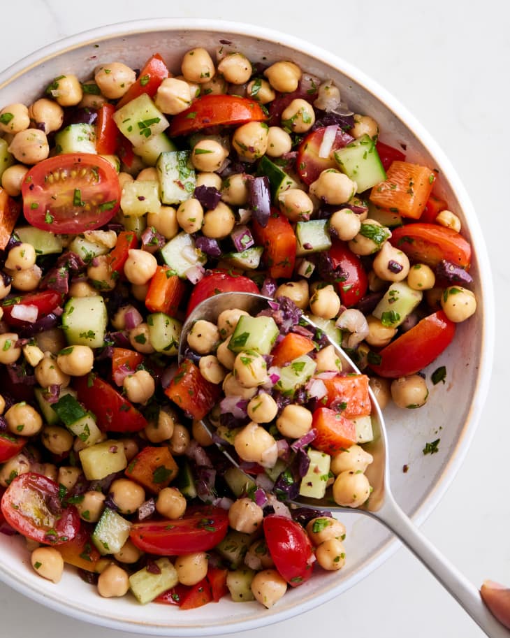 bowl of a mixed chickpea salad with chopped olives, red onions, red bell pepper,cucumber, and halved grape tomatoes on a marble surface with a scoop taken out with a silver serving spoon