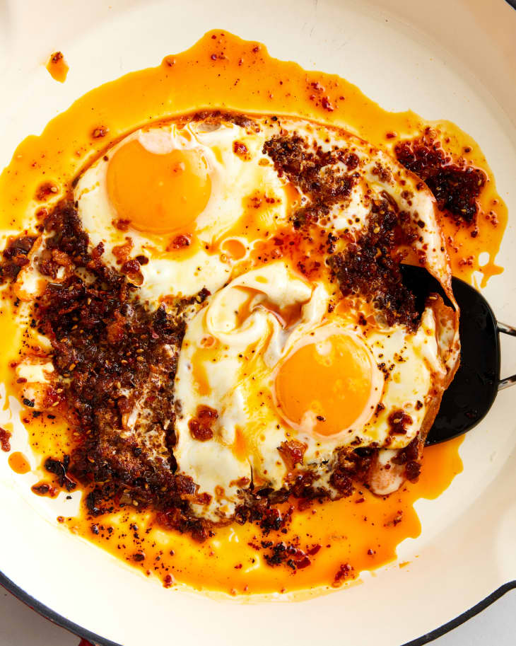 two eggs frying sunny side up over red chili crisp on a white skillet with a black spatula
