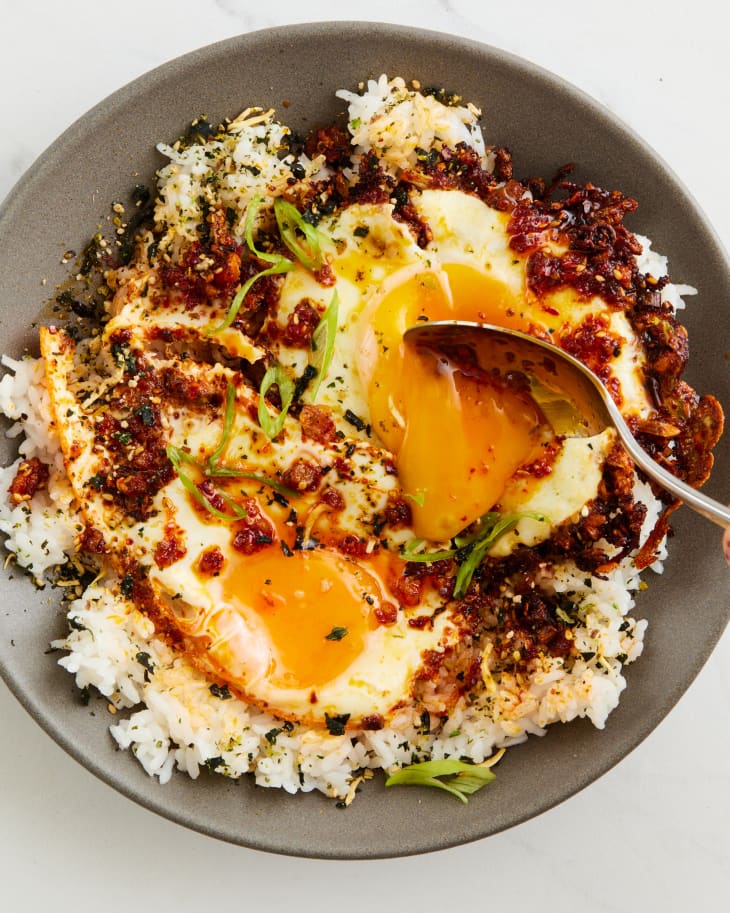 two fried sunny side up eggs with chili oil served over rice and garnished with scallions with one yolk broken and running