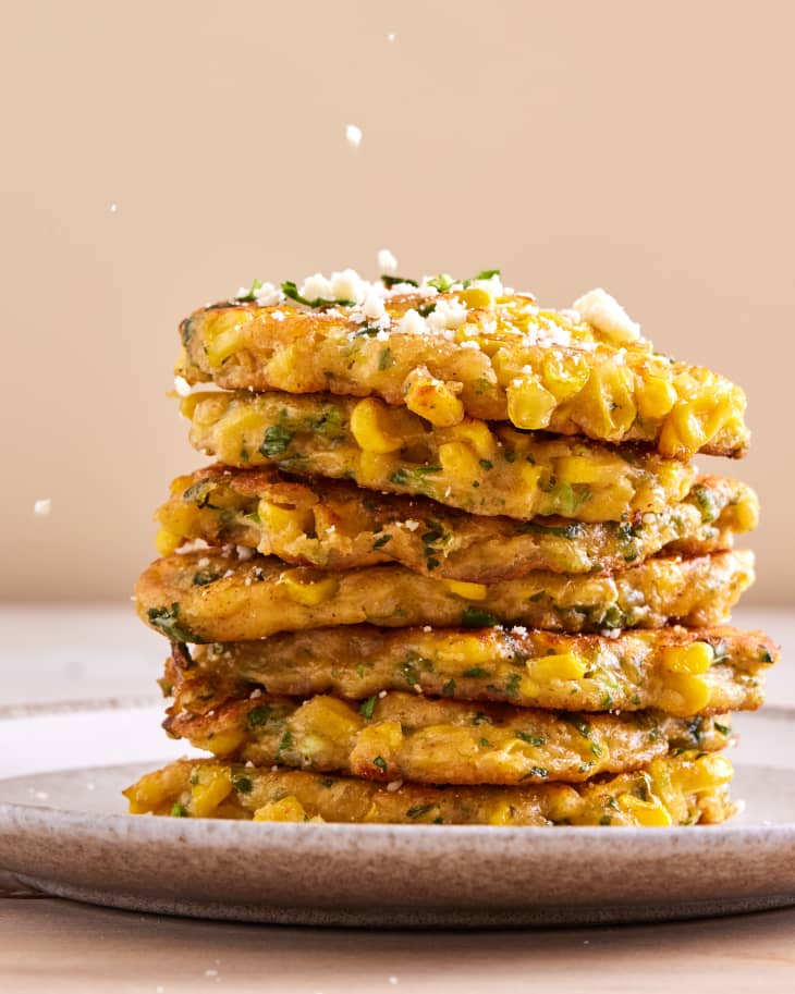 stack of seven elote(corn) fritters on a plate garnished with white cheese crumbles and chopped cilantro with additional cheese falling onto the stack