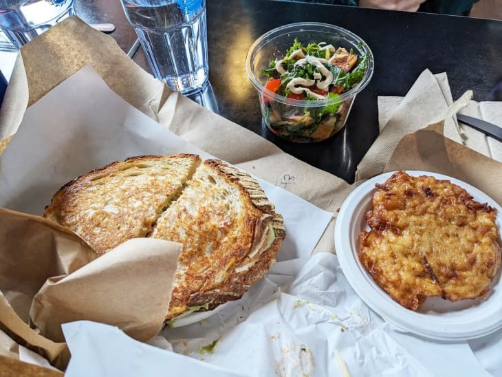 sandwich cut in half on parchment paper with side of potato pancakes and salad in a clear plastic cup