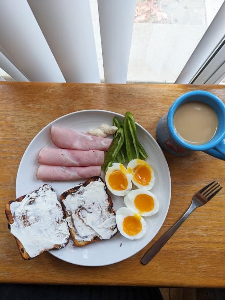 2 jammy eggs, toast with cream cheese, rolled pieces of ham, and pepper slices with a cup of coffee and a fork on the side