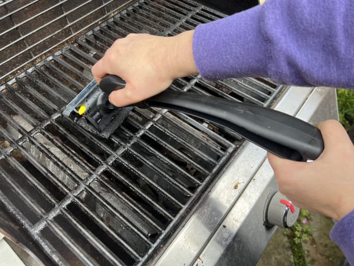 Hand scrubbing a barbecue grill grate with a black cleaning brush.