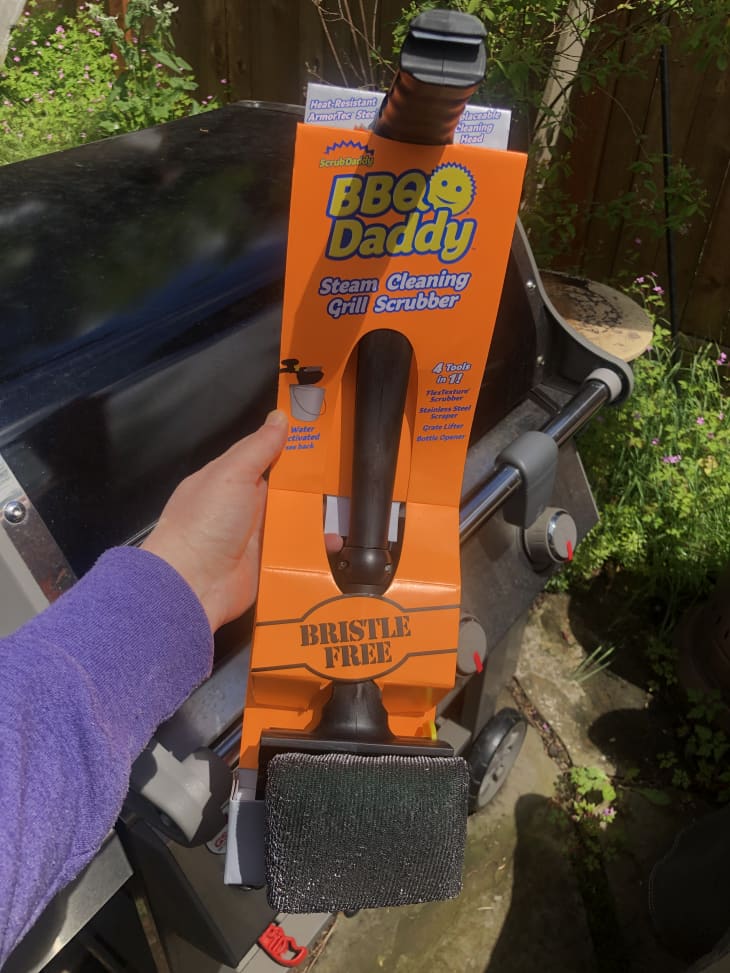 BBQ Daddy steam cleaning grill scrubber with bristle-free design held in front of a barbecue grill.