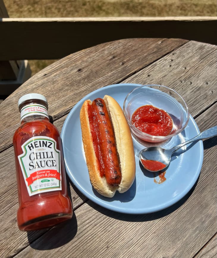plate with hot dog and chili sauce next to a bottle of chili sauce
