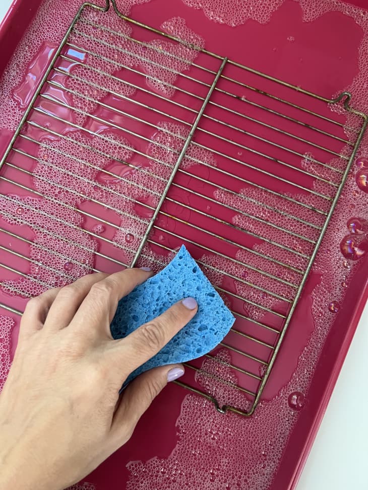 person using a sponge to clean a cooling rack on a baking sheet