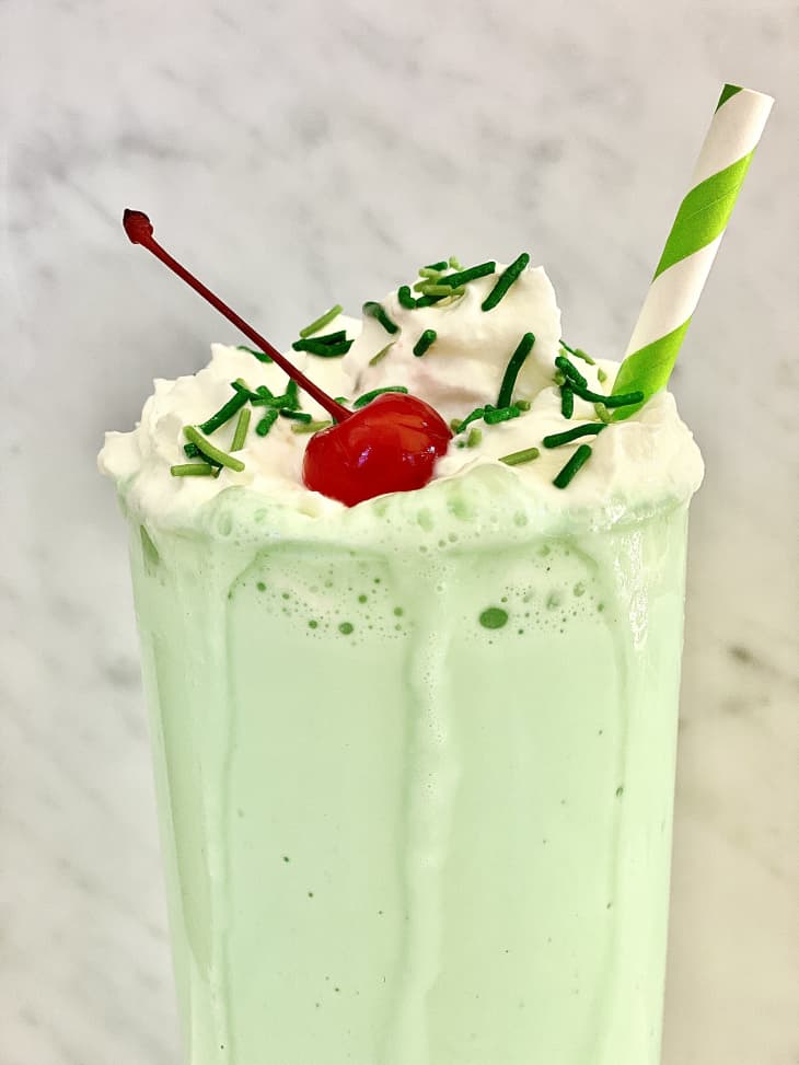 Shamrock shake up close with topped with whipped cream, green sprinkles, maraschino cherry and a green and white straw on the side.