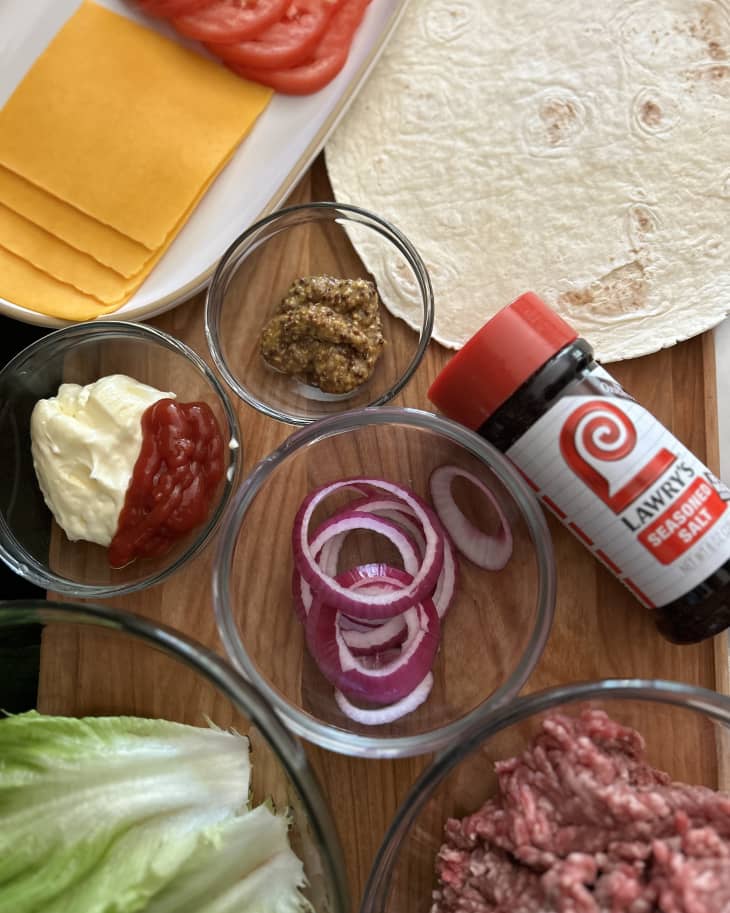 meal components for taco burger: sliced tomatoes, sliced cheese, red onions, Lawry's seasoned salt, lettuce, ground beef, tortillas, and condiments