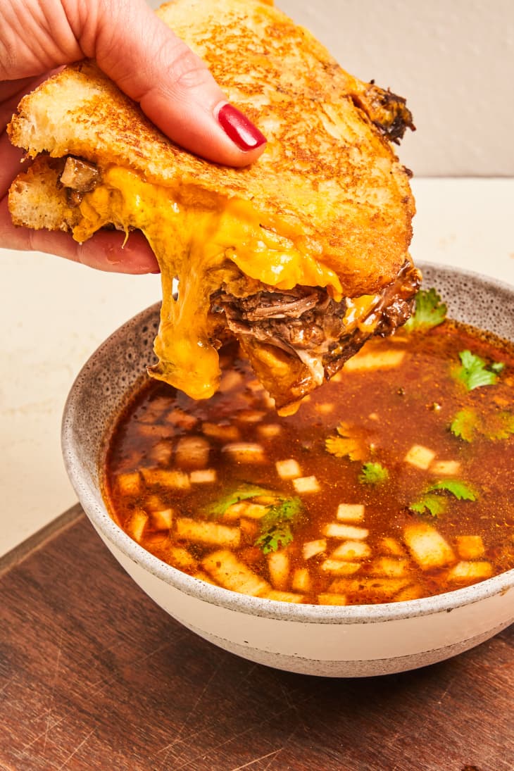 Someone dipping birria grilled cheese into consommmè.