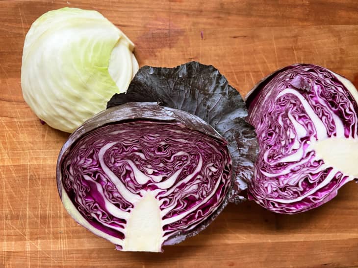 Red cabbage halved on cutting board with whole green cabbage behind.