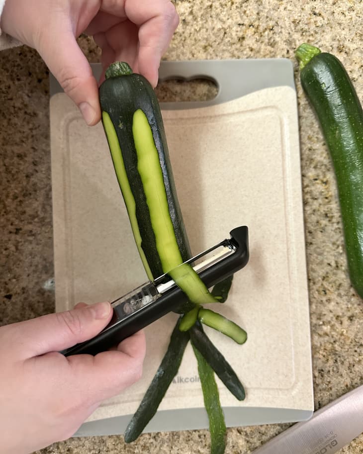 hands shown peeling zucchini squash with Dreamfarm Sharple peeler over cutting board